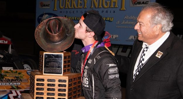 Logan Seavey kisses the Turkey Night Grand Prix trophy while J.C. Agajanian Jr. looks on. (Devin Mayo Photo)